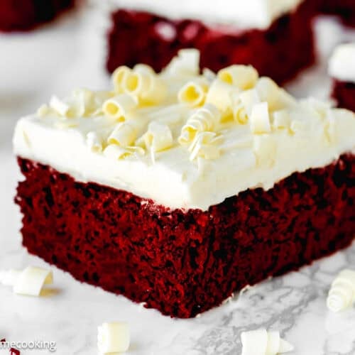 A square piece of eggless red velvet brownie topped with white frosting and white chocolate curls, placed on a marble surface.