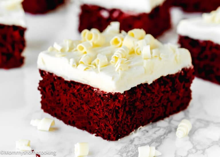 A square piece of eggless red velvet brownie topped with white frosting and white chocolate curls, placed on a marble surface.