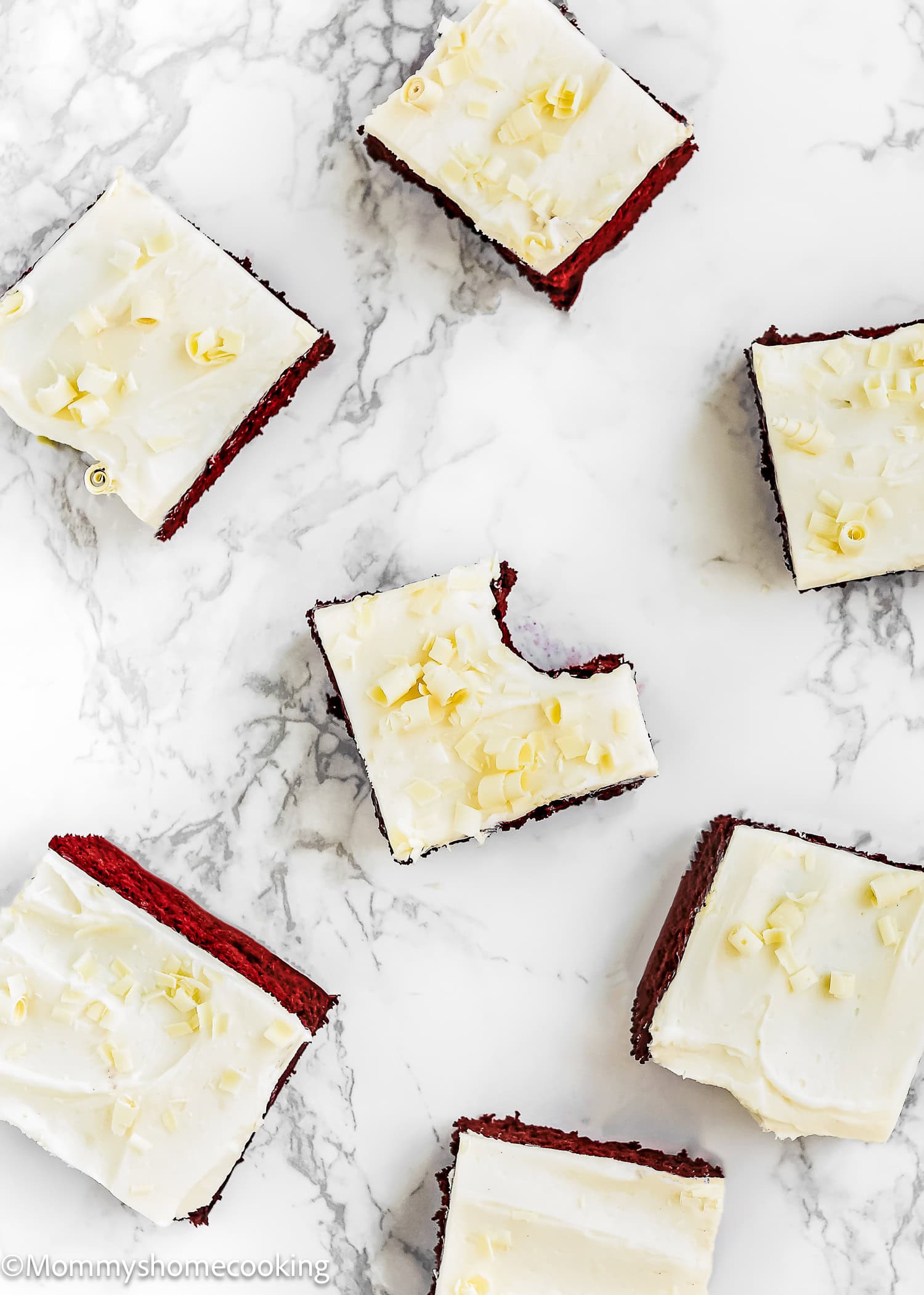 Seven frosted chocolate cake squares, some topped with white chocolate curls, are arranged on a marble surface. One piece has a bite taken out of it, showcasing the rich texture of Eggless Red Velvet Brownies.