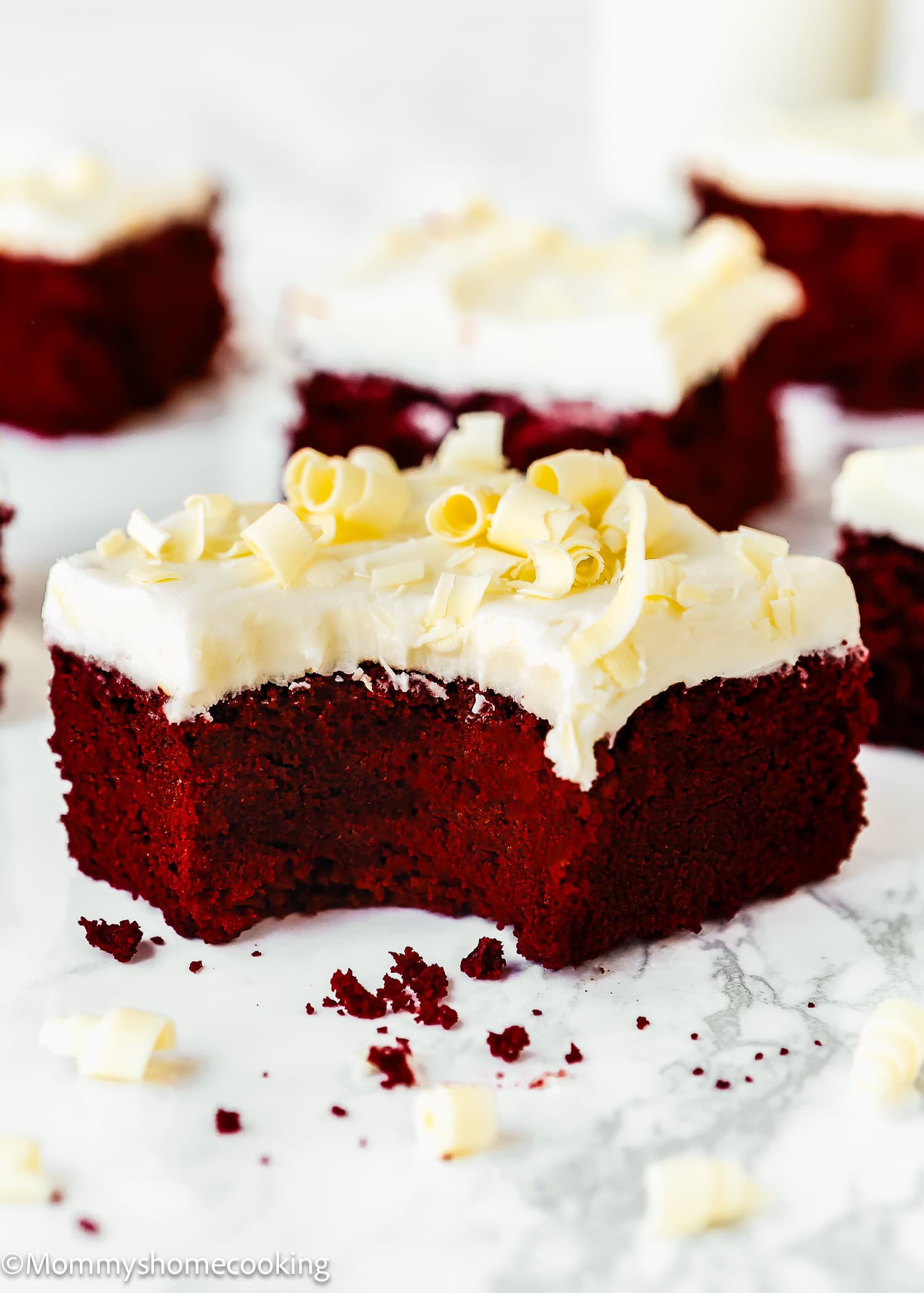 A piece of Eggless Red Velvet Brownies with white frosting and white chocolate curls, shown with a bite taken out, on a marble surface.