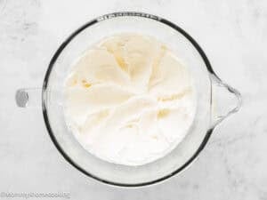 A glass mixing bowl filled with whipped cream sits on a white countertop, perfect for topping an Eggless Red Velvet Sheet Cake.
