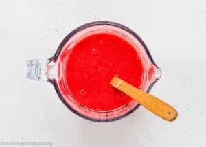 A glass measuring cup filled with red liquid, perfect for making Eggless Red Velvet Sheet Cake, and a wooden spoon rest on a white surface.