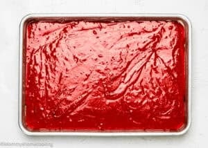 A rectangular baking sheet filled with a smooth layer of bright red liquid mixture, likely an Eggless Red Velvet Sheet Cake, sits on a white surface.
