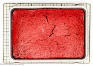 An Eggless Red Velvet Sheet Cake with cracks on the surface sits in a white baking dish on a wire cooling rack.