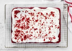 Eggless Red Velvet Sheet Cake with white frosting and red cake crumbs on top, placed on a wire rack lined with parchment paper.
