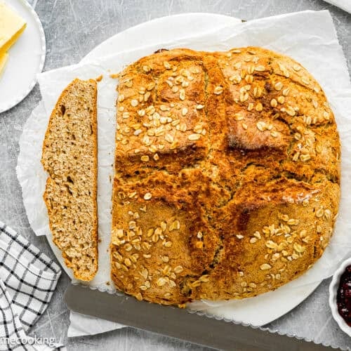 A round loaf of oat-topped Irish Soda Bread (No eggs!) with one slice cut, served on parchment paper next to butter, jam, a wooden knife, and a checkered cloth.