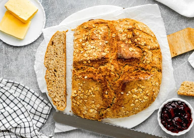 A round loaf of oat-topped Irish Soda Bread (No eggs!) with one slice cut, served on parchment paper next to butter, jam, a wooden knife, and a checkered cloth.
