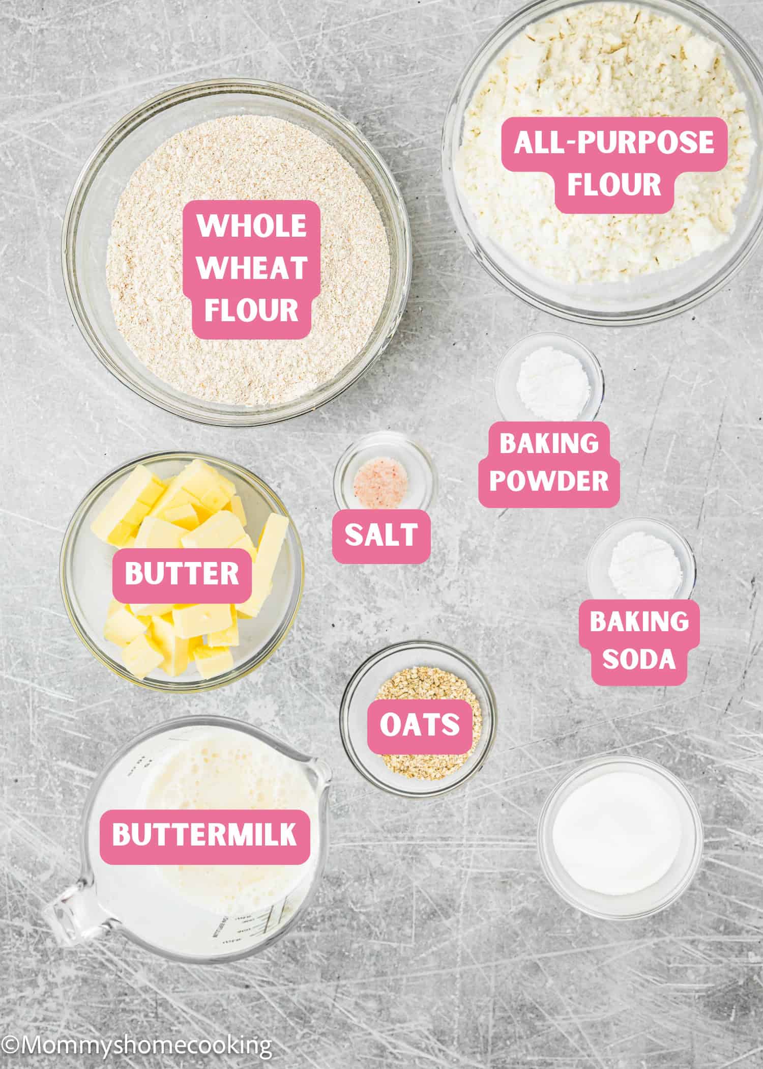 Top-down view of baking ingredients for Irish Soda Bread (No eggs!) in bowls labeled: whole wheat flour, all-purpose flour, butter, buttermilk, oats, salt, baking powder, and baking soda on a gray surface.