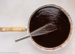 A metal whisk rests in a white saucepan filled with smooth, glossy chocolate sauce—perfect for making an Ultra-Fudgy Eggless Chocolate Layer Cake (Matilda Inspired)—on a light-colored surface.