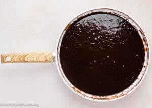 A white saucepan with a wooden handle filled with glossy, dark chocolate sauce sits on a light-colored surface—perfect for topping an Ultra-Fudgy Eggless Chocolate Layer Cake (Matilda Inspired).