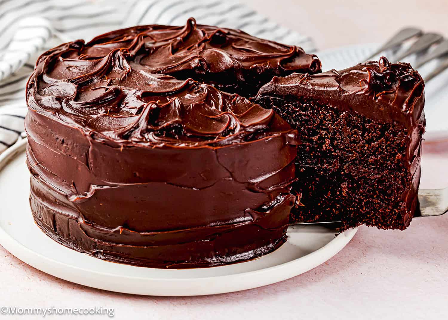 An ultra-fudgy eggless chocolate layer cake (Matilda inspired) with glossy chocolate frosting sits on a white plate, as one slice is lifted out on a spatula.