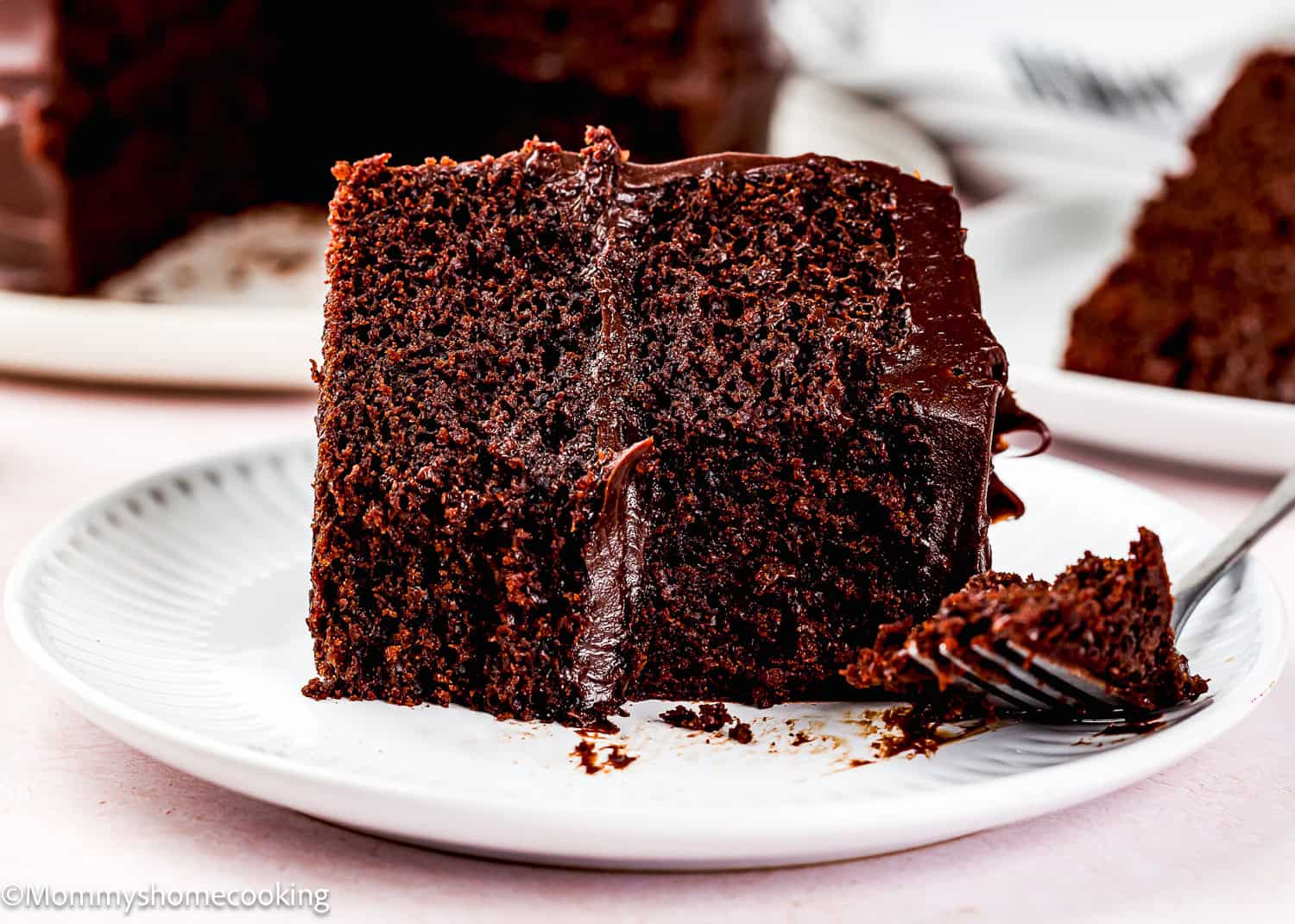 A slice of Ultra-Fudgy Eggless Chocolate Layer Cake (Matilda Inspired) with chocolate frosting sits on a white plate, a fork taking a bite.