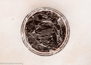 A glass bowl filled with chocolate pudding, covered tightly with plastic wrap, sits on a light-colored surface—perfect for pairing with an Ultra-Fudgy Eggless Chocolate Layer Cake (Matilda Inspired).