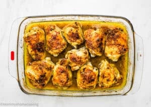 A glass baking dish filled with ten Easy Baked Lemon Chicken Thighs in a yellowish sauce on a white surface.