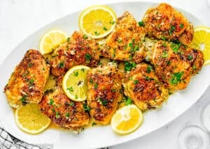 A white oval plate with Easy Baked Lemon Chicken Thighs garnished with parsley and lemon slices, set on a marble surface beside a fork, napkin, pepper grinder, and salt shaker.