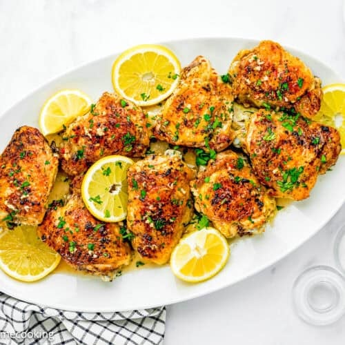 A white oval plate with Easy Baked Lemon Chicken Thighs garnished with parsley and lemon slices, set on a marble surface beside a fork, napkin, pepper grinder, and salt shaker.