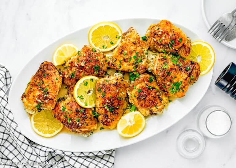 A white oval plate with Easy Baked Lemon Chicken Thighs garnished with parsley and lemon slices, set on a marble surface beside a fork, napkin, pepper grinder, and salt shaker.