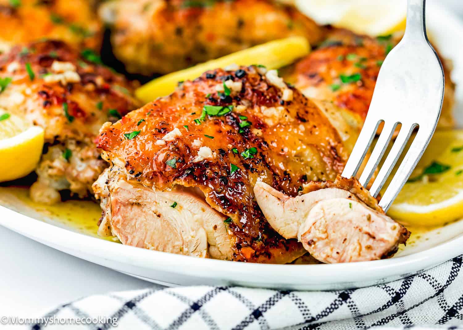 A close-up of Easy Baked Lemon Chicken Thighs garnished with herbs and lemon slices, with a fork cutting into a piece on a white plate.