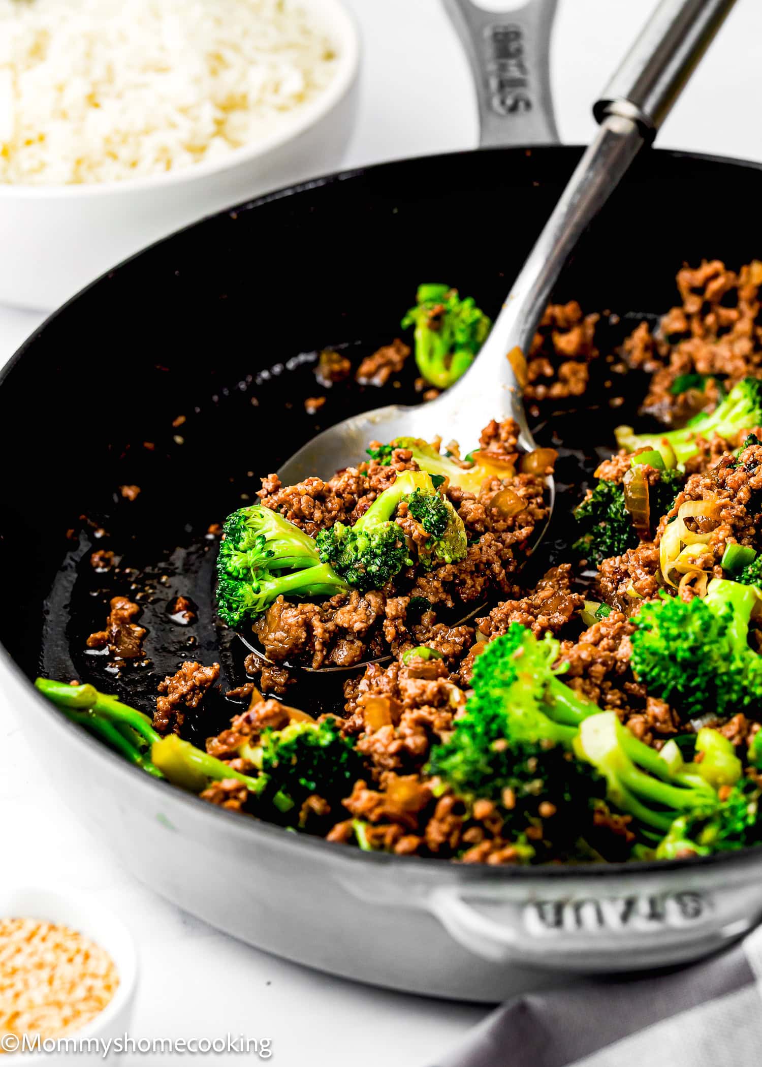 A skillet filled with Easy Ground Beef and Broccoli (20-Minute Dinner) and sauce is being served with a spoon; a bowl of white rice is visible in the background.