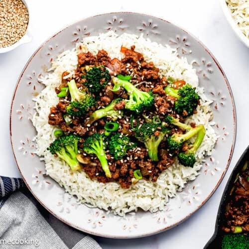 A plate of white rice topped with ground beef, broccoli, sliced green onions, and sesame seeds—an Easy Ground Beef and Broccoli (20-Minute Dinner) with extra bowls of rice and sesame seeds nearby.