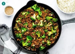 A skillet filled with cooked ground beef, broccoli, and onions next to a bowl of rice and a small dish of sesame seeds on a white surface—perfect for an Easy Ground Beef and Broccoli (20-Minute Dinner).