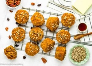 Twelve Egg-Free Morning Glory Muffins topped with sunflower seeds rest on a cooling rack, with pecans, raisins, honey, and butter arranged nearby on a white surface.