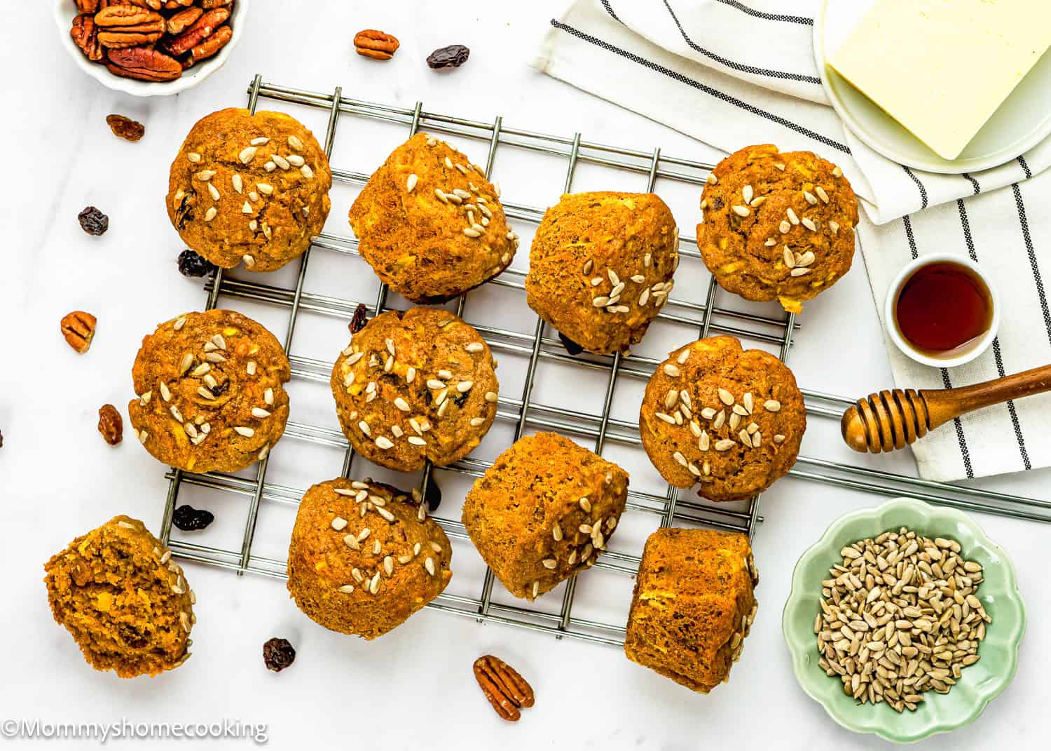Twelve Egg-Free Morning Glory Muffins topped with sunflower seeds rest on a cooling rack, with pecans, raisins, honey, and butter arranged nearby on a white surface.