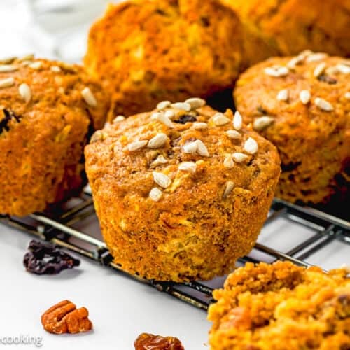 Egg-Free Morning Glory Muffins topped with sunflower seeds are cooling on a wire rack, with pecans and raisins scattered nearby.