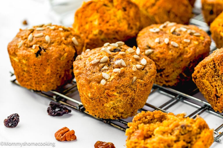 Egg-Free Morning Glory Muffins topped with sunflower seeds are cooling on a wire rack, with pecans and raisins scattered nearby.
