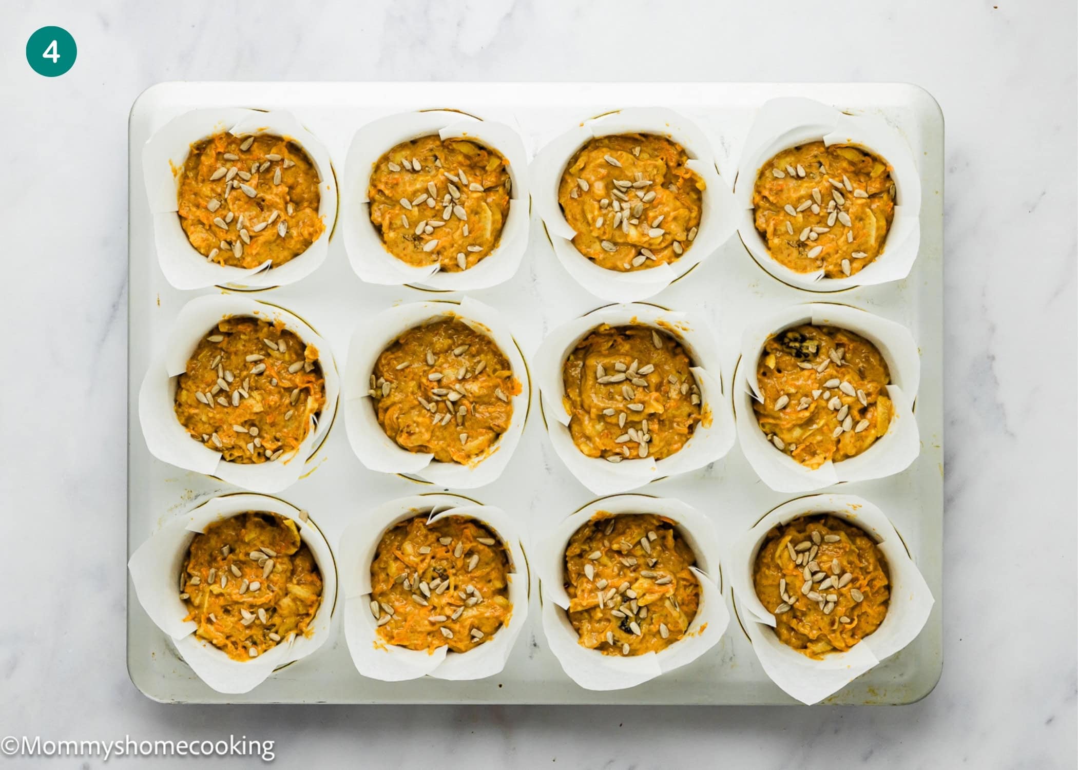 A muffin tray lined with white parchment paper holds raw Egg-Free Morning Glory Muffins batter topped with sunflower seeds, ready to be baked.