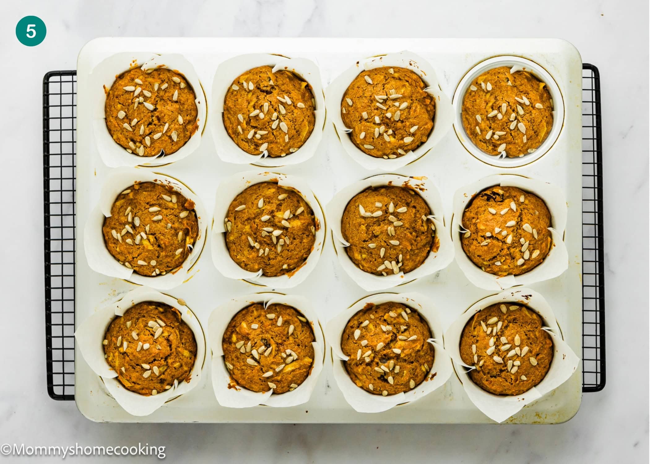 A muffin tray with twelve egg-free Morning Glory muffins topped with sunflower seeds, placed on a cooling rack over a white surface.
