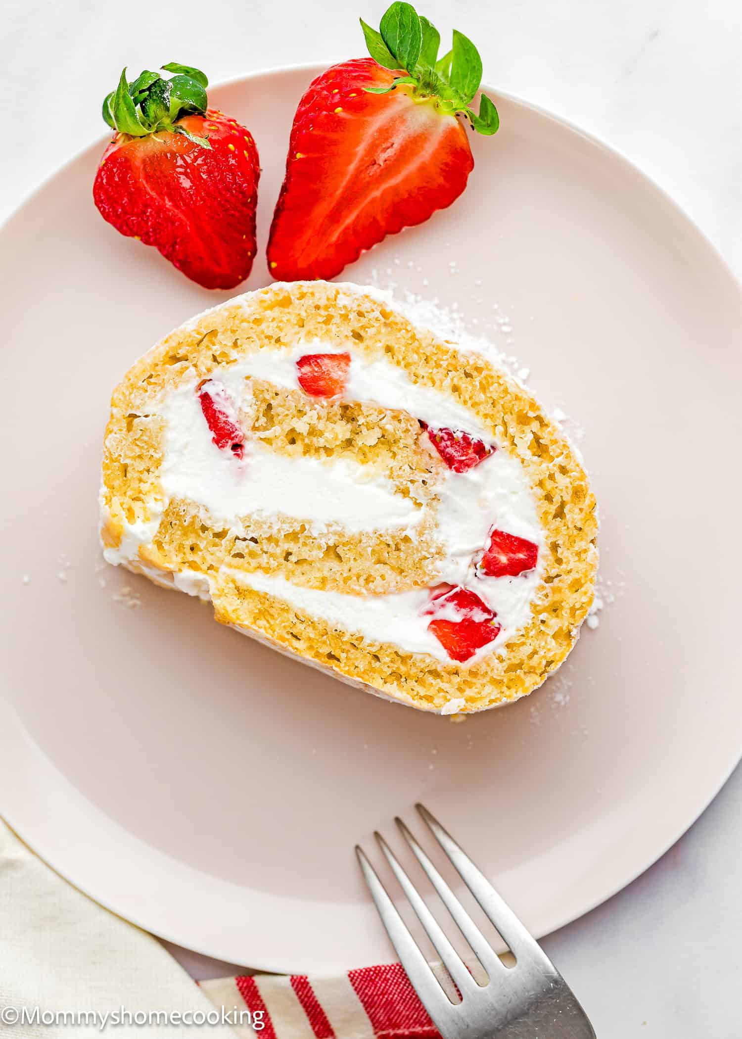 A slice of Egg-Free Vanilla Roll Cake filled with whipped cream and strawberries sits on a light pink plate, with two halved strawberries and a fork beside it.
