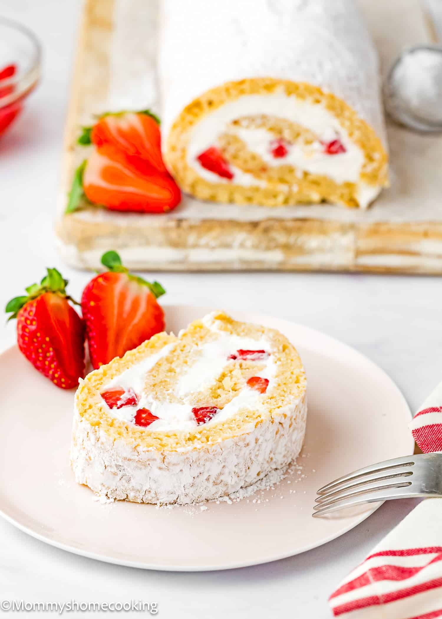 A slice of Egg-Free Vanilla Roll Cake with strawberries and cream sits on a plate, while fresh strawberries and the rest of the roll are displayed on a cutting board in the background.