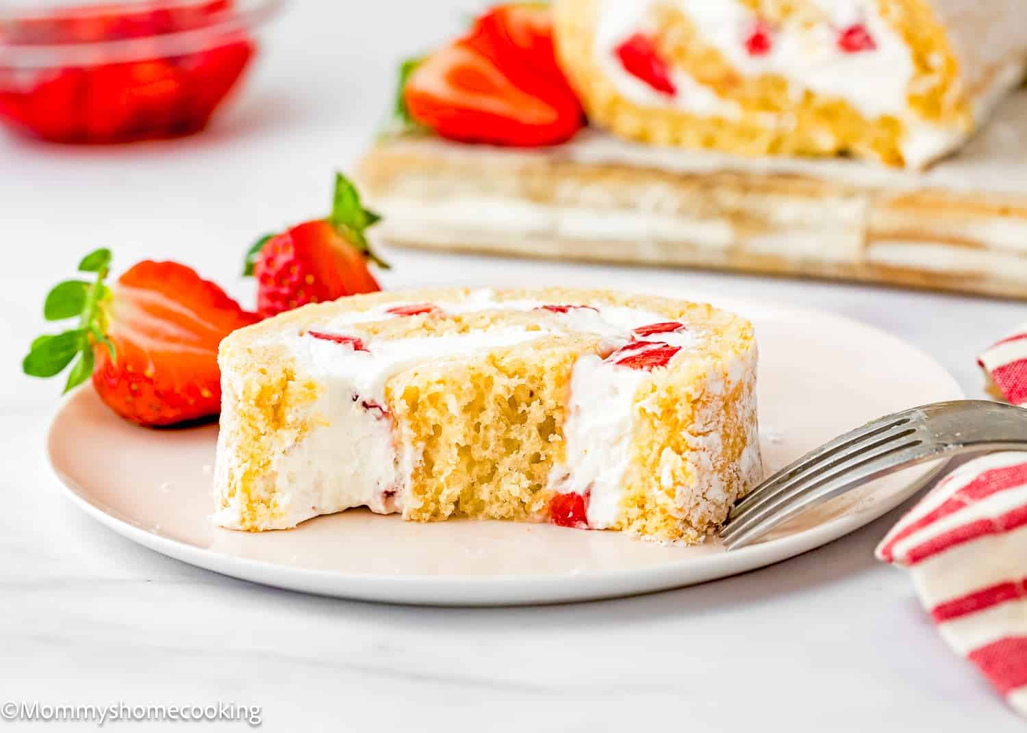 A slice of Egg-Free Vanilla Roll Cake filled with strawberries and cream on a plate with a fork, garnished with fresh strawberries.