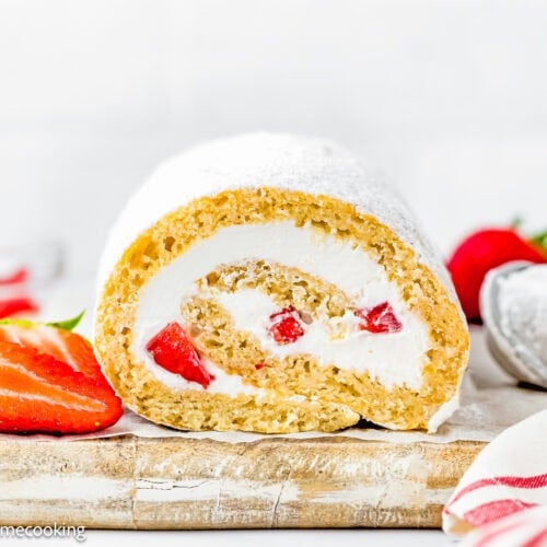 An Egg-Free Vanilla Roll Cake filled with strawberries and cream sits sliced on a wooden board, garnished with fresh strawberries, powdered sugar, and a red-striped cloth in the foreground.