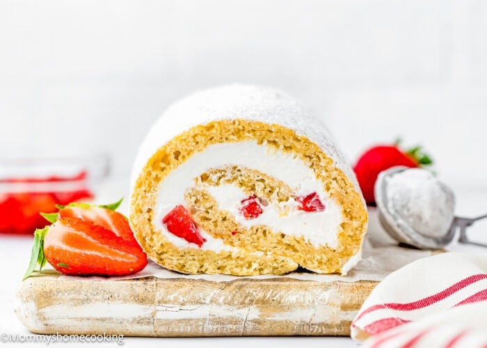 An Egg-Free Vanilla Roll Cake filled with strawberries and cream sits sliced on a wooden board, garnished with fresh strawberries, powdered sugar, and a red-striped cloth in the foreground.