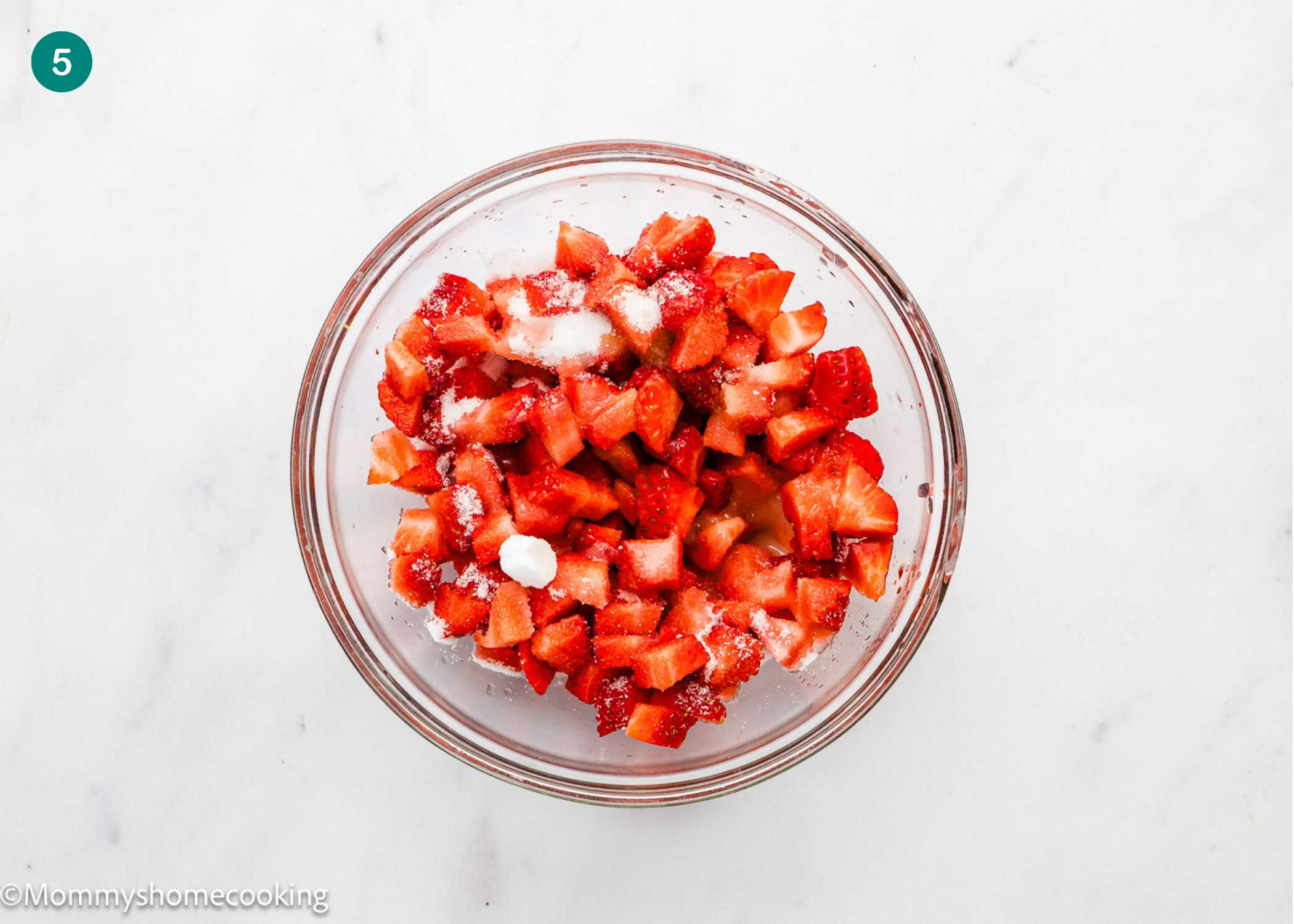 A glass bowl filled with chopped strawberries and sprinkled sugar sits on a white surface, perfect for pairing with an egg-free vanilla roll cake.