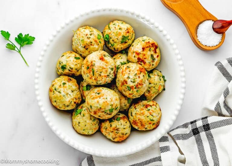 A white bowl filled with golden-brown Eggless Simple Air Fryer Turkey Meatballs (Juicy & Veggie Packed), garnished with chopped herbs, sits on a white surface beside a wooden salt holder, parsley, and a striped towel.