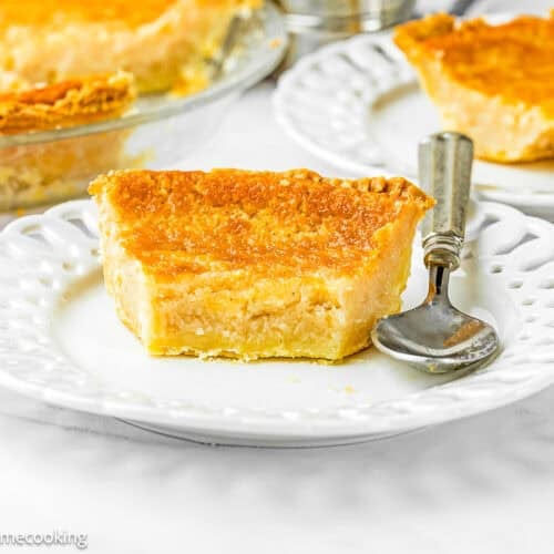 A slice of Eggless Buttermilk Pie on a white plate with a fork and spoon, with additional pie slices and a glass pie dish in the background.