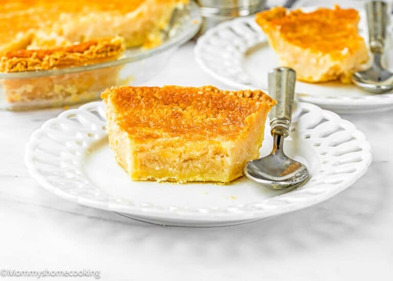 A slice of Eggless Buttermilk Pie on a white plate with a fork and spoon, with additional pie slices and a glass pie dish in the background.