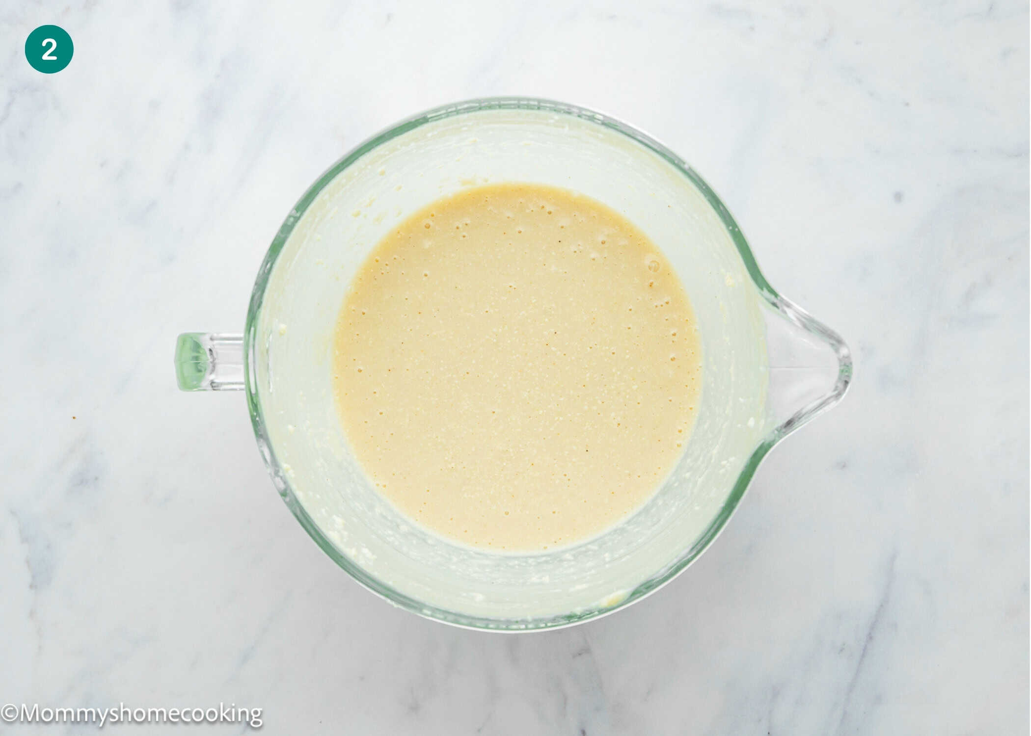 A clear glass mixing bowl filled with smooth, light yellow Eggless Buttermilk Pie batter sits on a white marble surface.