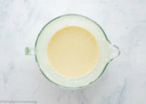 A glass mixing bowl filled with smooth, light yellow Eggless Buttermilk Pie batter sits on a white marble surface.