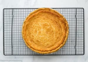 An Eggless Buttermilk Pie with a golden crust is cooling on a black wire rack placed on a white marble surface.