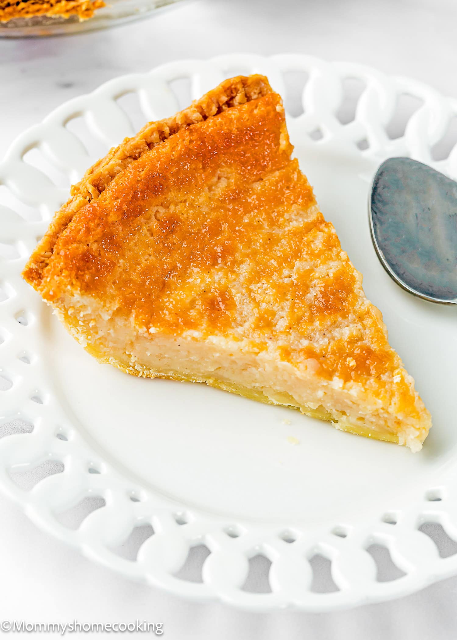 A single slice of Eggless Buttermilk Pie on a white decorative plate with a metal spoon beside it.
