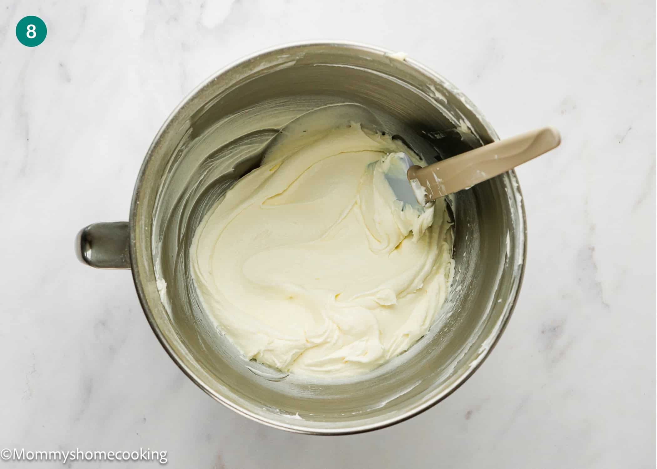 A mixing bowl with smooth, creamy frosting and a spatula resting inside, placed on a marble surface—perfect for finishing an Eggless Coconut Bundt Cake.