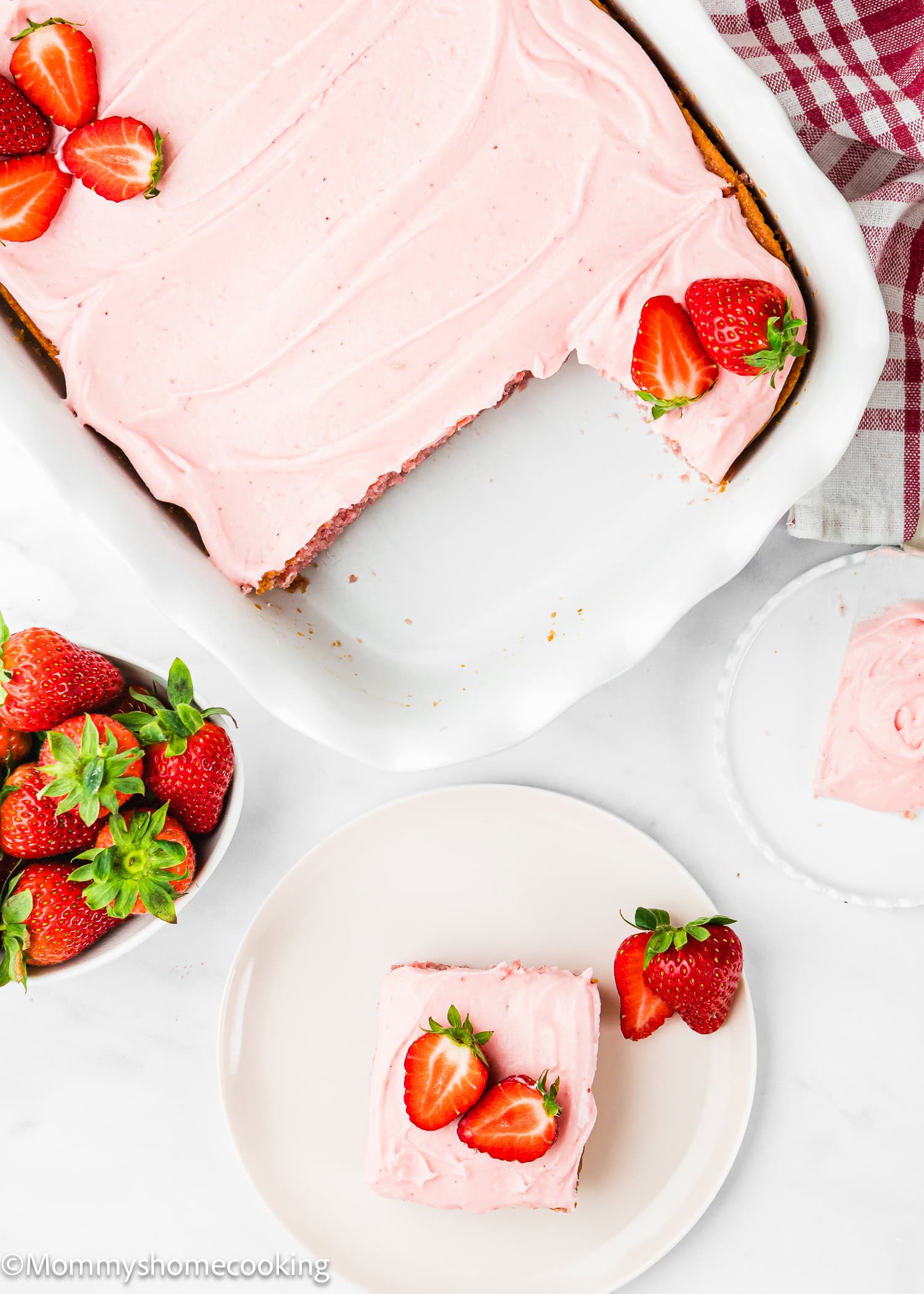 An Eggless Easy Strawberry Sheet Cake (Easy with Cake Mix) sits in a white dish with pink frosting, a slice on a plate, garnished with fresh strawberries. A bowl of strawberries is nearby.