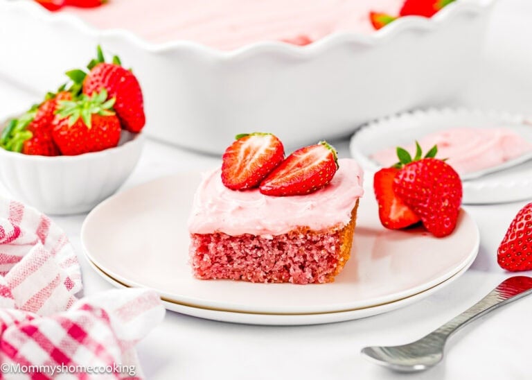 A slice of Eggless Easy Strawberry Sheet Cake with pink frosting and sliced strawberries on top sits on a white plate, with fresh strawberries and a baking dish in the background.