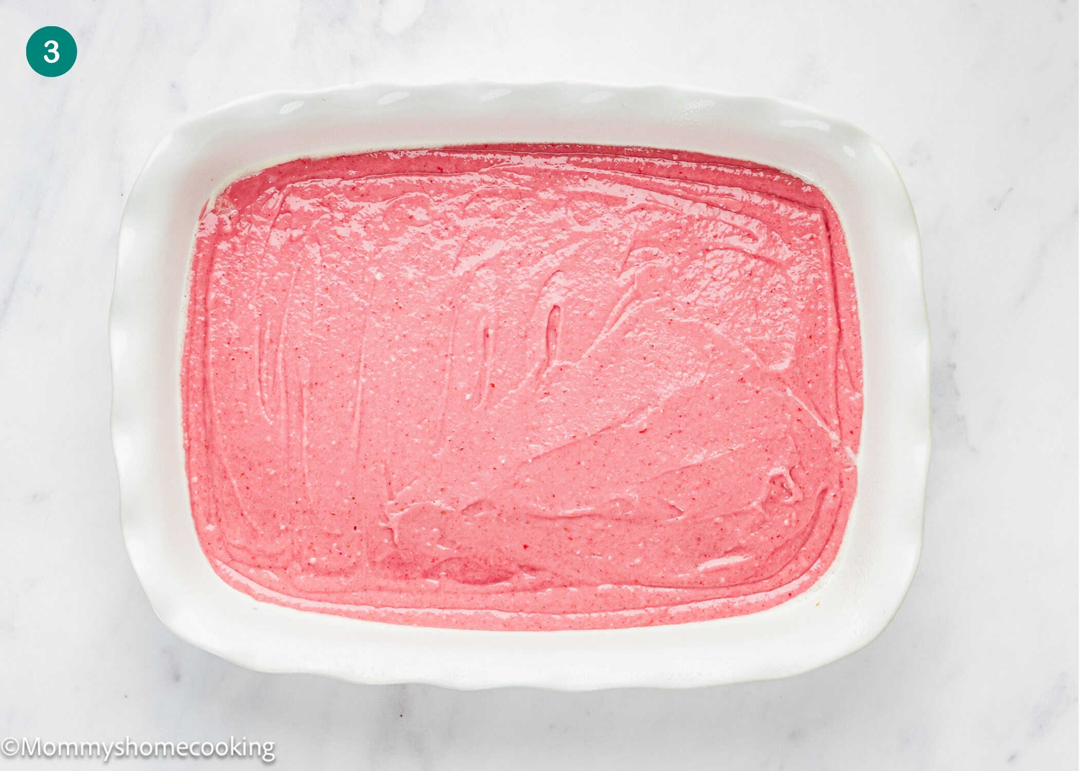 A white rectangular baking dish filled with smooth pink batter for an Eggless Easy Strawberry Sheet Cake (Easy with Cake Mix) sits on a white marble surface.