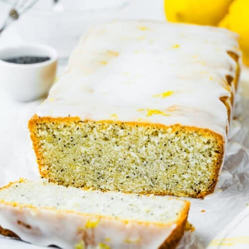 An Eggless Lemon Poppy Seed Loaf Cake with a sweet glaze, one slice cut, surrounded by fresh lemons and a small bowl of poppy seeds on a white background.