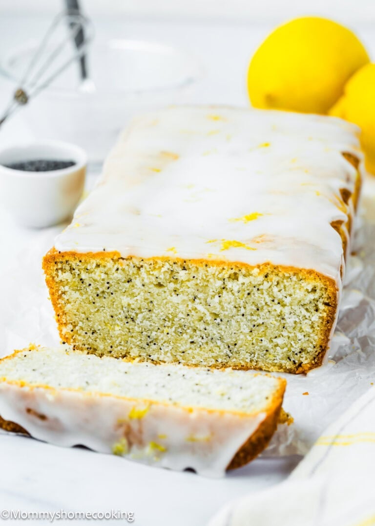 An Eggless Lemon Poppy Seed Loaf Cake with a sweet glaze, one slice cut, surrounded by fresh lemons and a small bowl of poppy seeds on a white background.